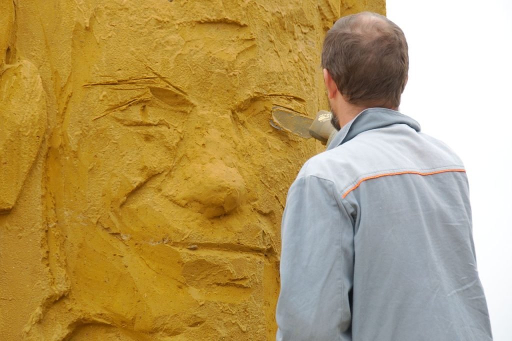 A skilled artist meticulously carves a detailed facial sculpture in sand, showcasing craftsmanship.